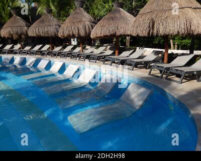 Area salotto piscina con ombrelloni di paglia, sedie a sdraio e palme paesaggio, in un resort caraibico sulla Riviera Maya, a Cancun, Mex Foto Stock