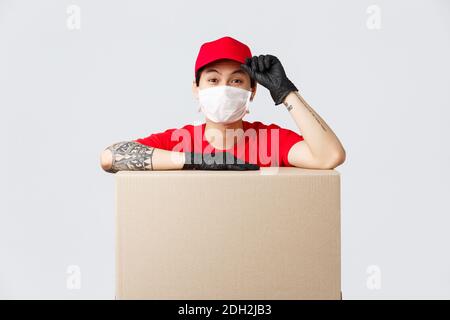 Ragazzo di consegna asiatico dall'aspetto amichevole in berretto rosso e t-shirt, servizio di corriere magra su scatola di cartone o pacchetto cliente, saluring As Foto Stock