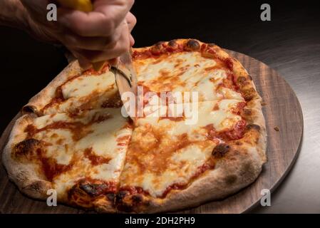 pizza con salsa di pomodoro e mozzarella, mano dello chef con fette di coltello a rullo su tagliere di legno Foto Stock