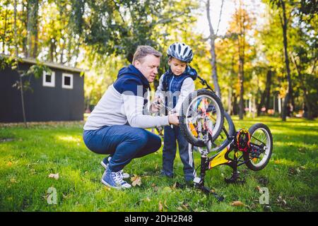 Papà caucasico e figlio di 5 anni nel cortile vicino alla casa sul prato verde riparando una bicicletta, Foto Stock