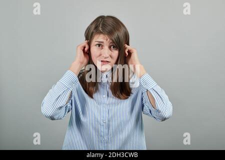 L'inserimento delle orecchie con le dita non vuole sentire, mani coperte dell'auricolo, dispiaciuto, blocca il suono, qualcosa è forte. Giovane donna attraente, camicia blu vestita con occhi marroni, capelli brunette, grigio Foto Stock