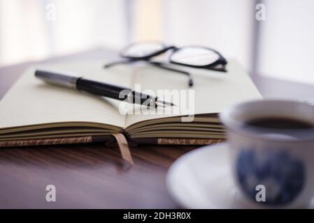 Un colpo di fuoco selettivo di una tazza di caffè, taccuino, penna e occhiali Foto Stock
