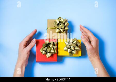 Le mani della donna che prendono o gioiscono tre regali colorati, rossi, gialli e artigianali, con nastri di lusso dorati su sfondo blu lignt. Foto Stock