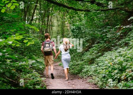 Felice coppia caucasica stanno camminando in una fitta foresta lungo il percorso tenendo le mani, vista posteriore. Escursionisti con zaino alla ricerca di p Foto Stock