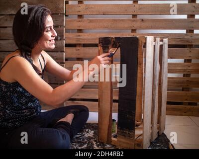 Giovane donna ispanica sorridente vestita con abiti scuri dipinge A. Scatola di legno in nero Foto Stock