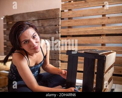 Giovane donna ispanica sorridente vestita con abiti scuri dipinge A. Scatola di legno in nero e guarda la fotocamera Foto Stock