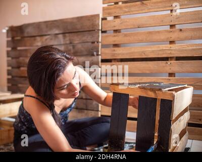 Giovane donna ispanica vestita in vestiti scuri dipinge una scatola di legno in nero, con uno sfondo di pallet Foto Stock