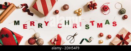 Allegra iscrizione natalizia fatta con lettere decorative ad albero, scatole regalo festive, carta da imballaggio, baubles, nastri su sfondo bianco, vista dall'alto. Biglietto d'auguri alla moda per le vacanze di Natale, striscione Foto Stock