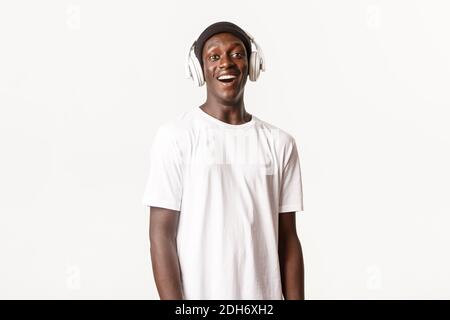 Ritratto di allegro e felice afro-americano giovane ragazzo ascoltando podcast o musica in cuffia, sorridente felice, bianco backgro Foto Stock