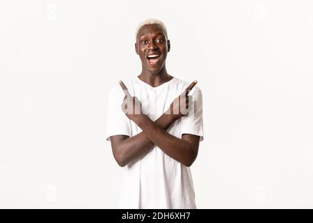 Ritratto di felice ed eccitato, attraente biondo afroamericano, puntando le dita lateralmente e sorridendo stupito, backgr bianco Foto Stock