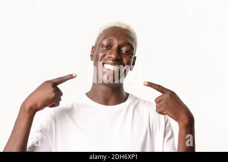 Primo piano di felice bel ragazzo biondo afroamericano che punta al suo sorriso bianco perfetto, consiglia dentista, essere felice, sta Foto Stock