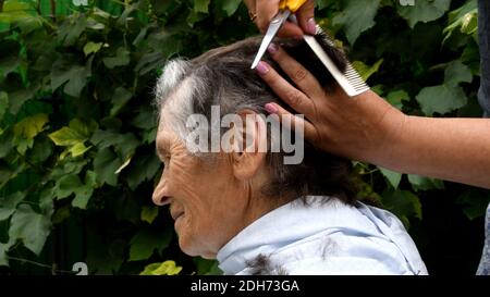 Taglio dei capelli di donna anziana a casa in cortile esterno. Le mani femminili tengono il pettine e le forbici per tagliare i capelli corti della donna di 80 anni Foto Stock
