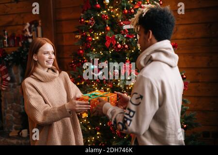 Giovane afroamericano che dà il regalo di Natale alla moglie caucasica sull'albero di natale di sfondo. Foto Stock