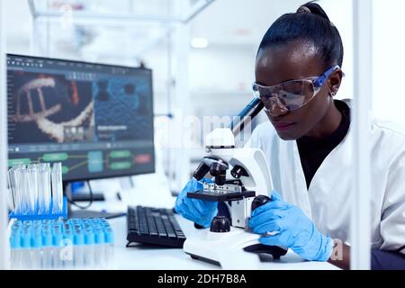 Virologo di etnia africana seduto sul suo posto di lavoro tubi di prova puliti guardando attraverso il microscopio. Scienziato sanitario nero in laboratorio di biochimica che indossa attrezzature sterili. Foto Stock