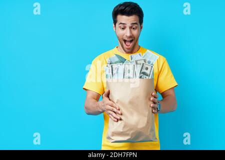 Eccitato felice uomo tenere sacchetto di carta pieno di pile di denaro isolato su sfondo blu. Foto Stock