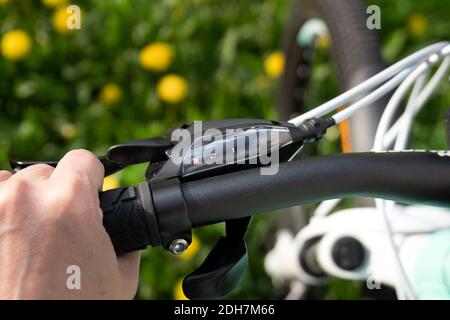 Selettore del cambio di bicicletta sul manubrio con la mano che tiene una maniglia e i diandelioni gialli sull'erba verde sullo sfondo sfocato in primavera. Foto Stock