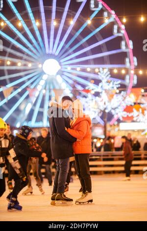 Uomo e donna innamorati in abbraccio su arena di ghiaccio contro di ruota Ferris in serata di Natale. Coppia alla pista di pattinaggio di Roma Foto Stock