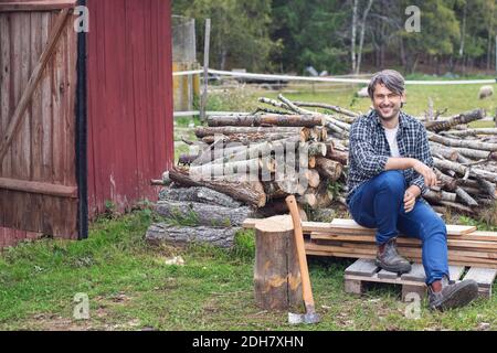 Ritratto di uomo seduto su tavole di legno contro legna da ardere accatastata in cantiere Foto Stock