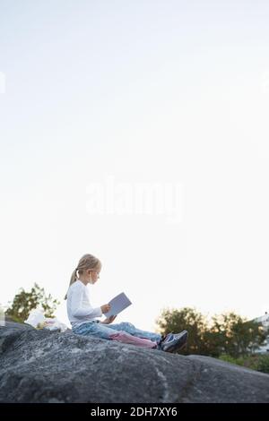 Vista laterale completa della ragazza che fa i compiti mentre si siede sulla roccia contro il cielo limpido Foto Stock