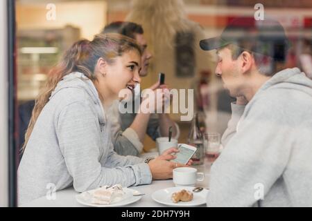 Visualizzazione di una giovane coppia che parla mentre si utilizza lo smartphone in caffè attraverso il vetro Foto Stock