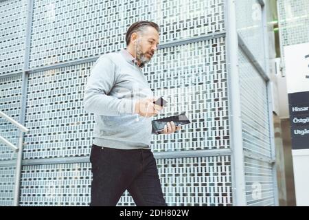 Uomo d'affari con tablet digitale che ascolta musica tramite telefono cellulare mentre camminare in ufficio Foto Stock