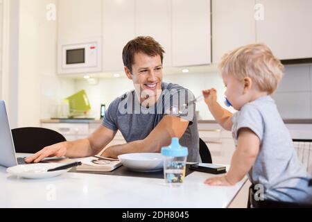 Buon padre che guarda il bambino mentre usa il computer portatile cucina Foto Stock