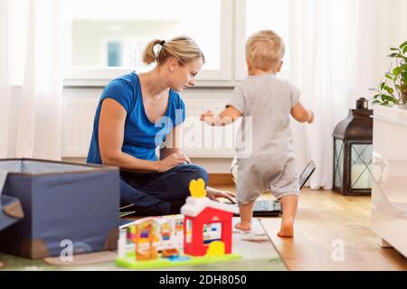 Bambino che cammina verso la madre usando il computer portatile a casa Foto Stock