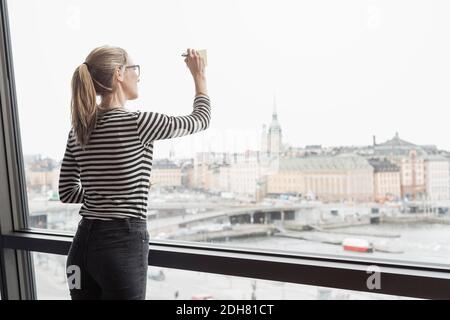 Vista posteriore di una donna d'affari che scrive sulla nota adesiva attaccata a. vetrata in ufficio Foto Stock