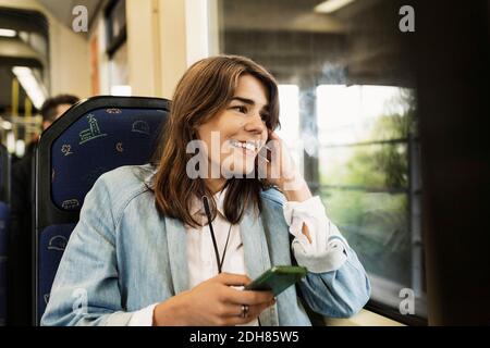 Sorridente giovane donna che tiene il telefono cellulare mentre si guarda attraverso il tram finestra Foto Stock