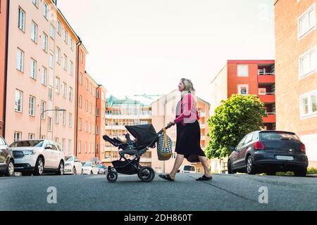 Vista laterale completa della donna che spinge il bambino nel carrello attraversando la strada della città Foto Stock