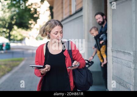 Donna mid adulta che usa il telefono cellulare mentre si lascia per lavoro con la famiglia in background Foto Stock