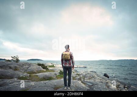 Vista posteriore a tutta lunghezza di un uomo in piedi sulla roccia mare contro cielo nuvoloso Foto Stock