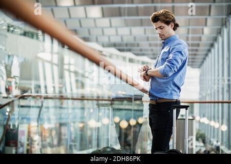 Vista laterale di un uomo d'affari che controlla il tempo mentre si è in piedi ringhiera in vetro all'aeroporto Foto Stock