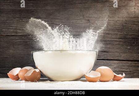 farina bianca che fuoriesce da una ciotola di vetro e dalle gusci di uovo su sfondo rustico Foto Stock