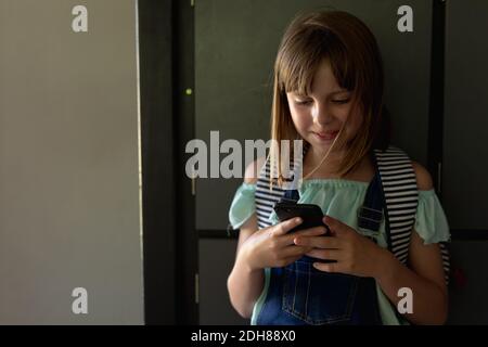 Schoolgirl appoggiato contro gli armadietti in un corridoio utilizzando uno smartphone Foto Stock