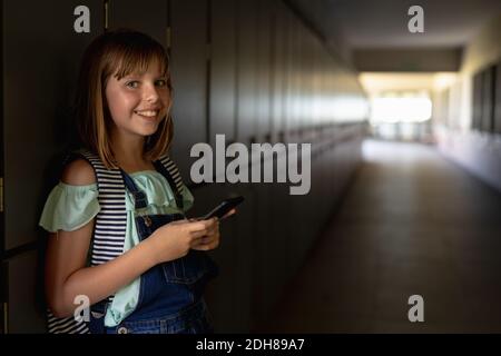 Schoolgirl appoggiato contro gli armadietti in un corridoio utilizzando uno smartphone Foto Stock