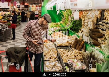 Uomo che tiene il telefono intelligente mentre comprando la verdura di radice nel supermercato Foto Stock