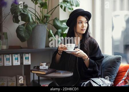 Donna giovane premurosa che tiene la tazza di caffè mentre si siede vicino alla finestra in ufficio creativo Foto Stock