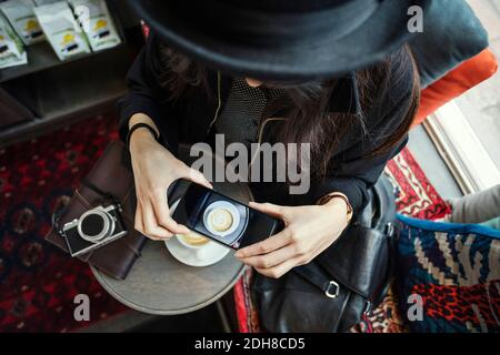 Direttamente sopra il colpo di giovane donna che fotografa il caffè attraverso Smart telefono in ufficio creativo Foto Stock