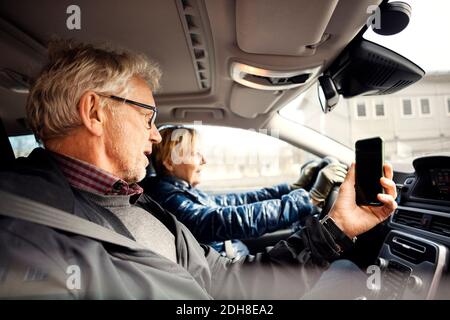 Uomo che tiene lo smartphone mentre si siede da una donna anziana che guida auto in città Foto Stock