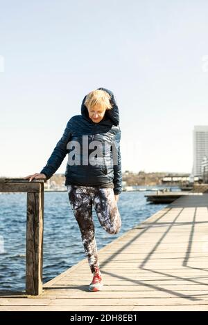 Donna anziana che allunga la gamba mentre si sta sul molo contro chiaro cielo Foto Stock