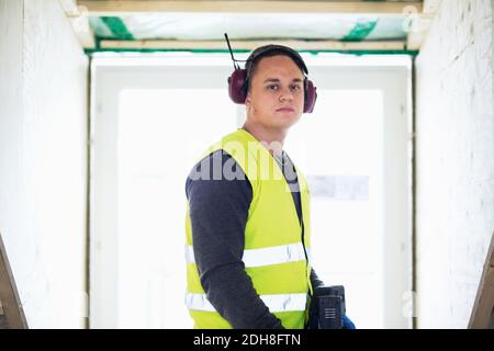 Ritratto di un uomo serio che indossa le cuffie protettive in piedi contro la finestra Foto Stock