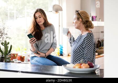 Madre che beve caffè mentre guarda la figlia usando lo smartphone in cucina Foto Stock