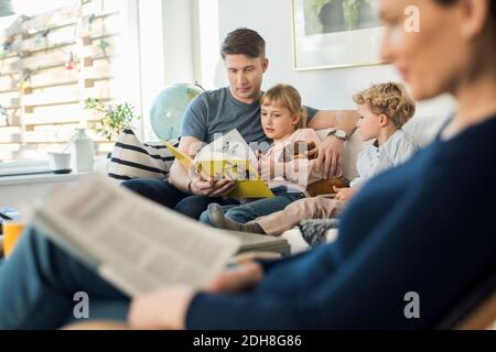 Padre leggere libro di storia per i bambini con la madre seduta dentro soggiorno Foto Stock