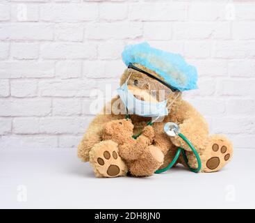 l'orso bruno del teddy si siede in occhiali protettivi di plastica, una maschera medica usa e getta e un cappuccio blu, concetto di pediatria Foto Stock