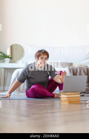 Donna anziana che ha video conversazione con un addestratore in linea prima di un workout Pilates. Lei dice ciao e sventolando la sua mano nella fotocamera del laptop. Concetto Foto Stock