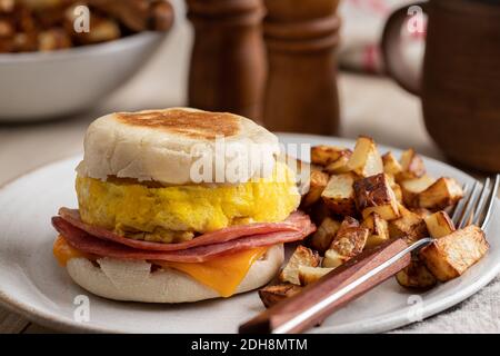 Sandwich per la colazione su un muffin inglese fatto con uova, prosciutto e formaggio con patate fritte su un piatto Foto Stock