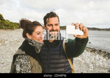 Felice coppia che prende selfie alla spiaggia contro il cielo durante il tramonto Foto Stock