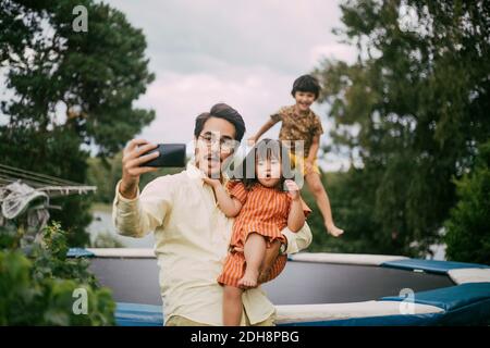 Sorridente padre che porta giù la figlia di sindrome mentre prendendo selfie contro ragazza che salta sul trampolino durante l'estate Foto Stock