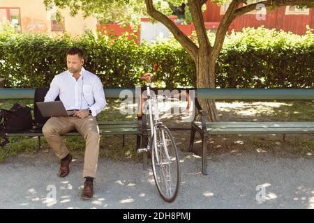 Uomo d'affari che usa il computer portatile mentre si siede sulla panca del parcheggio Foto Stock